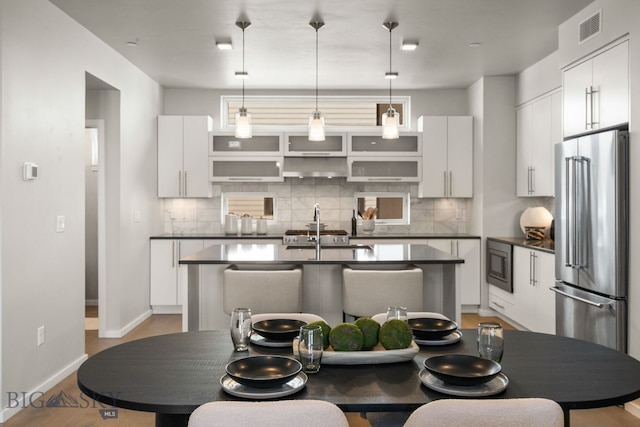 kitchen with white cabinetry, hanging light fixtures, a kitchen island with sink, stainless steel appliances, and decorative backsplash