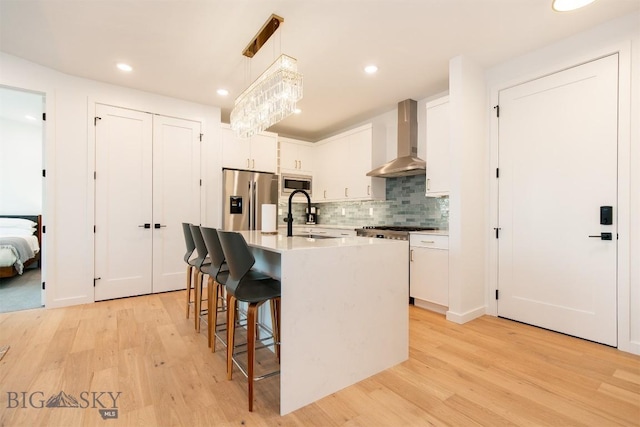 kitchen featuring pendant lighting, white cabinetry, stainless steel appliances, an island with sink, and wall chimney exhaust hood