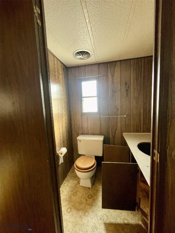 half bath with visible vents, toilet, vanity, wooden walls, and a textured ceiling