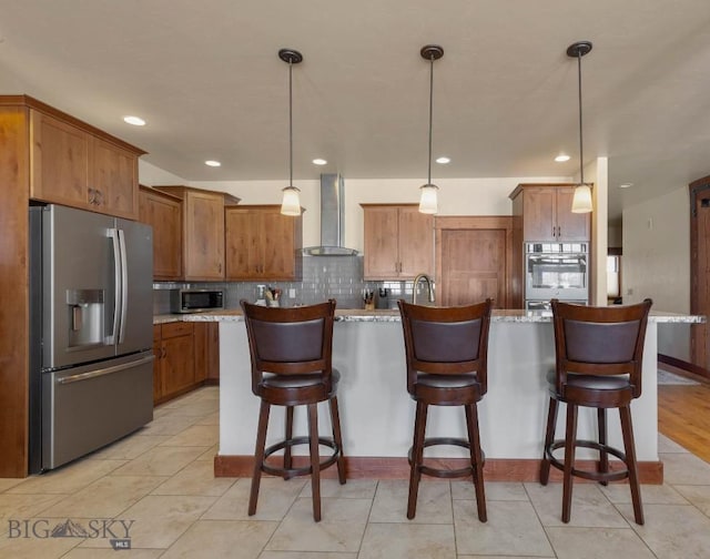kitchen with a kitchen island with sink, wall chimney range hood, hanging light fixtures, and appliances with stainless steel finishes