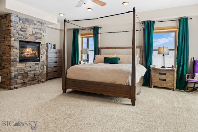 carpeted bedroom featuring a stone fireplace, a ceiling fan, multiple windows, and recessed lighting