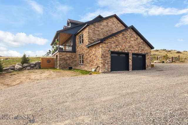view of side of home with an attached garage, gravel driveway, stone siding, and a balcony