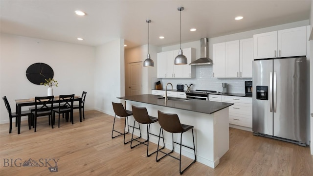 kitchen featuring stainless steel appliances, dark countertops, a kitchen island with sink, white cabinetry, and wall chimney exhaust hood