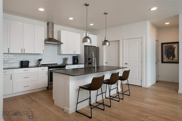 kitchen featuring a center island with sink, stainless steel appliances, dark countertops, white cabinetry, and wall chimney exhaust hood