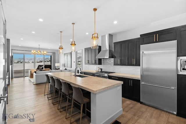 kitchen with built in appliances, dark cabinets, light countertops, wall chimney range hood, and an island with sink