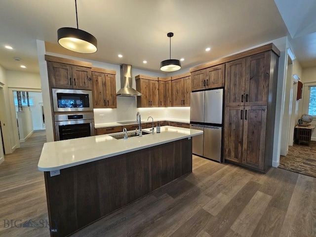 kitchen with stainless steel appliances, light countertops, hanging light fixtures, a kitchen island with sink, and wall chimney range hood