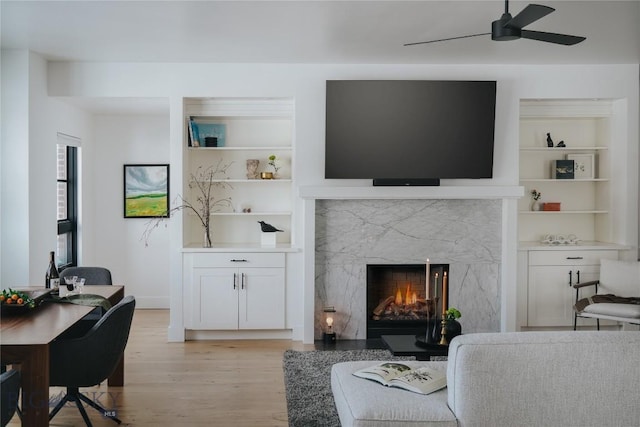 living area featuring light wood-type flooring, built in shelves, ceiling fan, and a premium fireplace