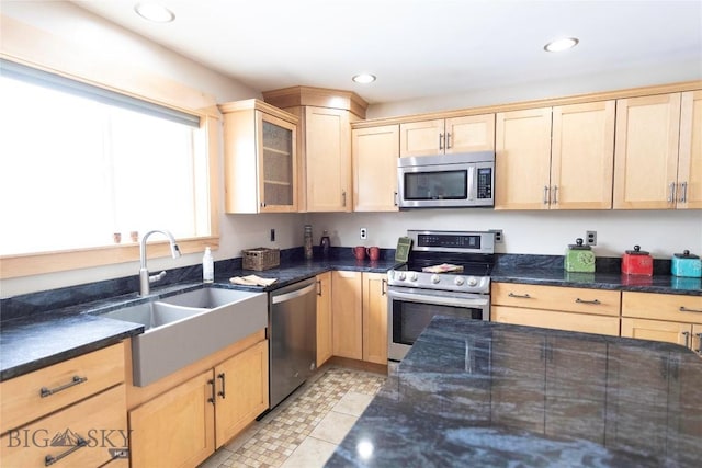 kitchen with light brown cabinets, stainless steel appliances, a sink, dark stone countertops, and glass insert cabinets