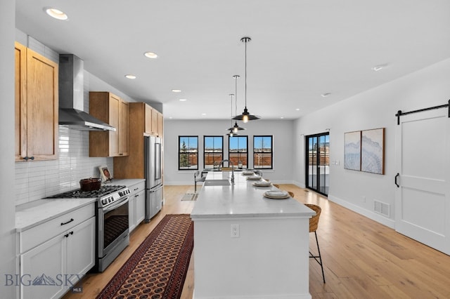 kitchen with a barn door, visible vents, an island with sink, stainless steel appliances, and wall chimney range hood