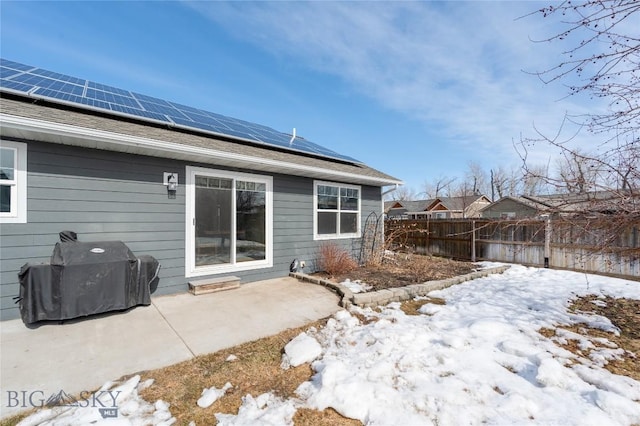 snow covered house featuring a shingled roof, roof mounted solar panels, a patio area, and fence