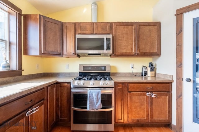 kitchen featuring light wood finished floors, stainless steel appliances, light countertops, brown cabinetry, and vaulted ceiling