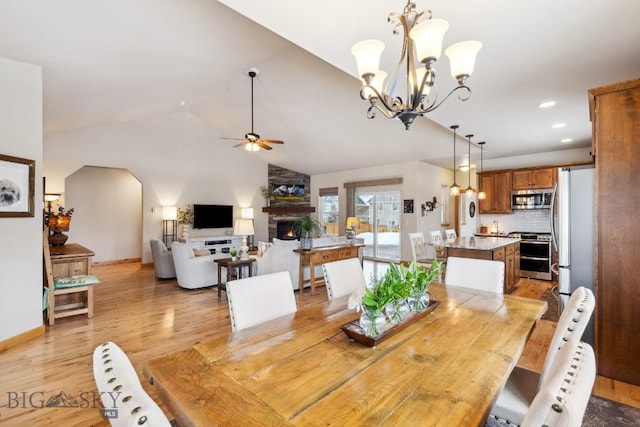 dining area featuring light wood-style flooring, ceiling fan with notable chandelier, recessed lighting, a stone fireplace, and vaulted ceiling