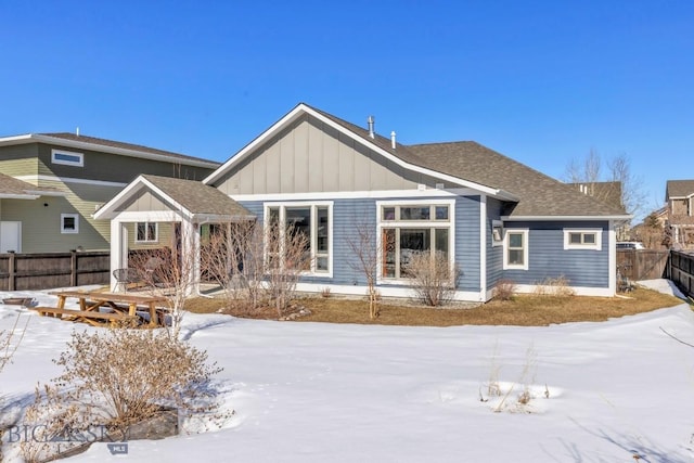 snow covered house with board and batten siding, roof with shingles, and fence