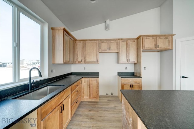 kitchen with light brown cabinets, a sink, dark stone counters, light wood finished floors, and lofted ceiling