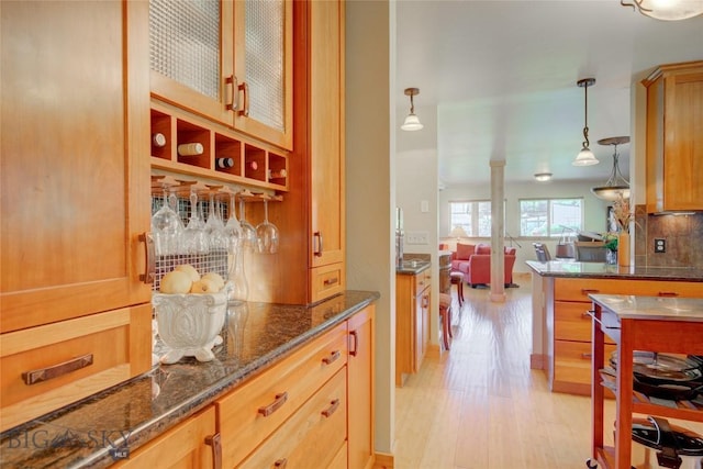 kitchen featuring light wood finished floors, glass insert cabinets, decorative light fixtures, decorative backsplash, and dark stone countertops