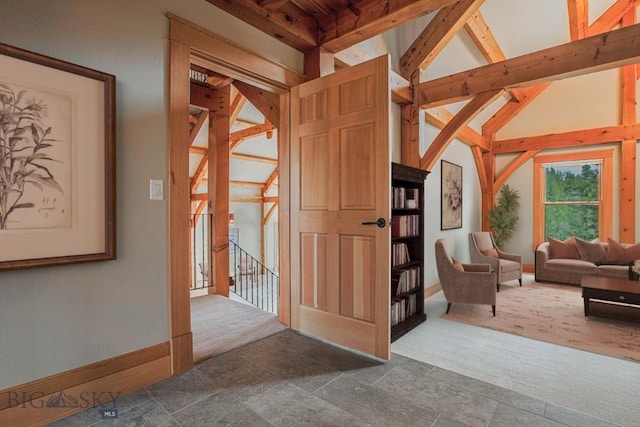 corridor with an upstairs landing, stone finish floor, dark colored carpet, baseboards, and vaulted ceiling with beams