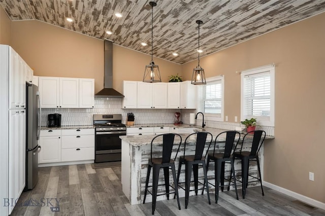 kitchen featuring wall chimney range hood, light stone counters, appliances with stainless steel finishes, a peninsula, and white cabinetry