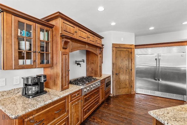 kitchen with brown cabinetry, built in appliances, dark wood finished floors, and glass insert cabinets