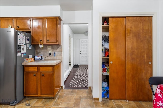 kitchen with tile countertops, brown cabinetry, freestanding refrigerator, stone finish floor, and tasteful backsplash