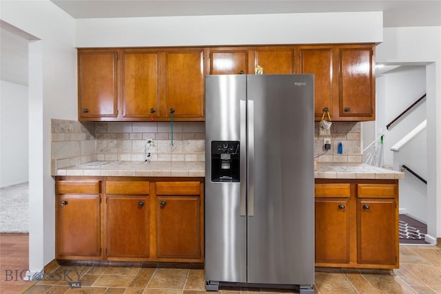 kitchen featuring brown cabinetry, tile countertops, stainless steel fridge with ice dispenser, and backsplash