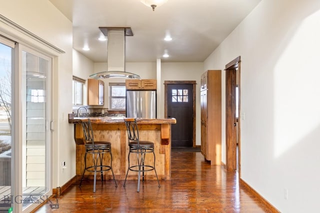 kitchen featuring dark wood finished floors, a peninsula, stainless steel refrigerator, a kitchen breakfast bar, and island range hood