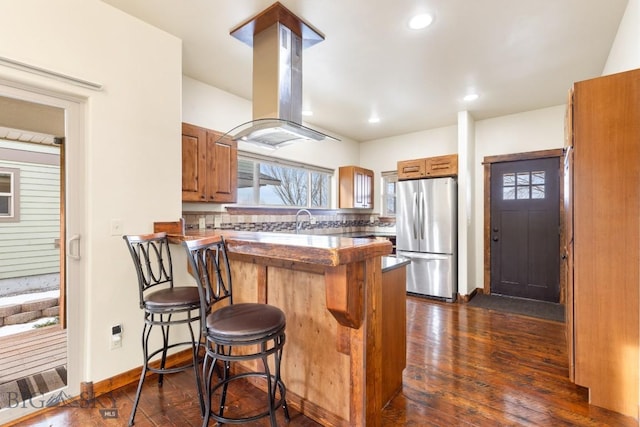 kitchen featuring brown cabinetry, a peninsula, island exhaust hood, freestanding refrigerator, and a kitchen breakfast bar