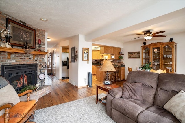 living room featuring wood finished floors, baseboards, ceiling fan, a textured ceiling, and a brick fireplace