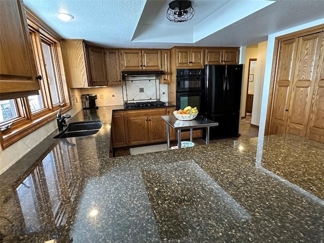 kitchen featuring dark stone counters, a sink, black appliances, under cabinet range hood, and a textured ceiling