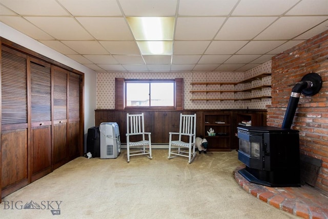 living area featuring a paneled ceiling, light carpet, wood walls, and a wood stove