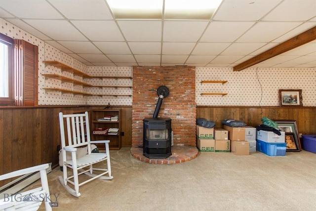 interior space featuring a wood stove, carpet, a drop ceiling, wooden walls, and wainscoting