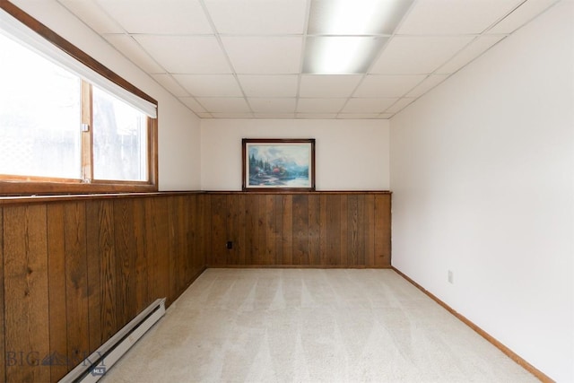 empty room with wood walls, baseboard heating, a drop ceiling, light carpet, and wainscoting