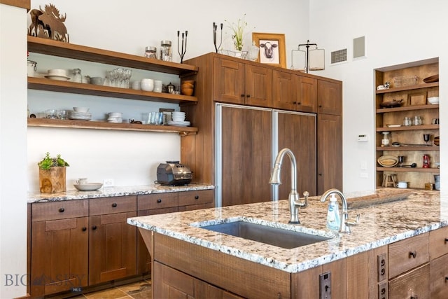 kitchen featuring a sink, paneled built in fridge, open shelves, brown cabinetry, and light stone countertops