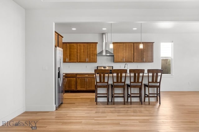 1511 Masterson, Belgrade, MT 59714 - kitchen featuring a breakfast bar area, brown cabinetry, light wood-type flooring, stainless steel refrigerator with ice dispenser, and wall chimney exhaust hood