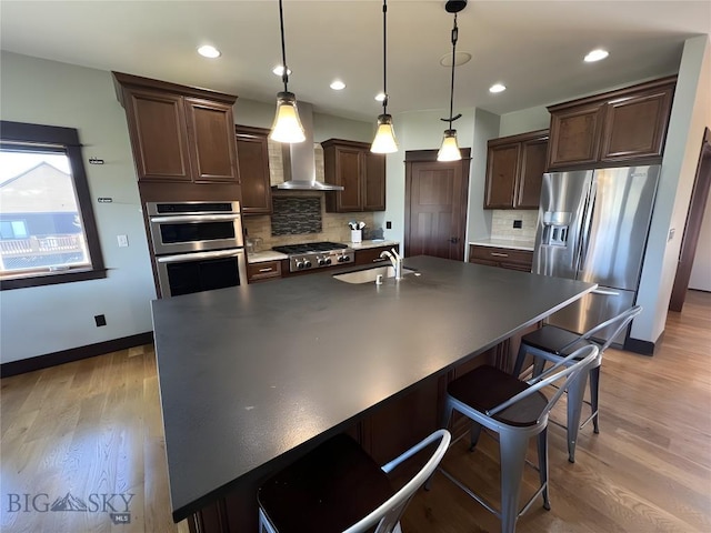 kitchen with wall chimney range hood, light wood-style flooring, a sink, stainless steel appliances, and dark brown cabinets