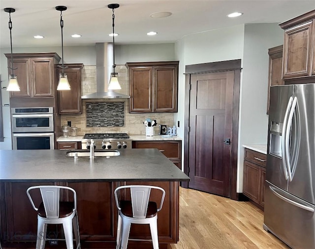 kitchen featuring a sink, light wood-style floors, wall chimney exhaust hood, a breakfast bar area, and appliances with stainless steel finishes