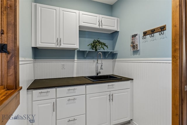 kitchen featuring white cabinetry, dark countertops, a sink, and a wainscoted wall