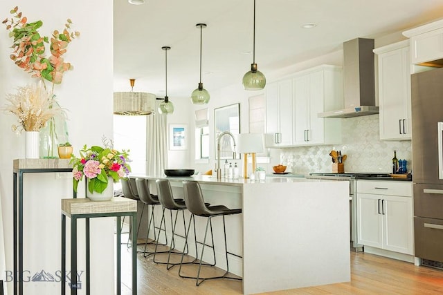kitchen featuring light wood-style flooring, wall chimney exhaust hood, white cabinetry, decorative backsplash, and a center island with sink