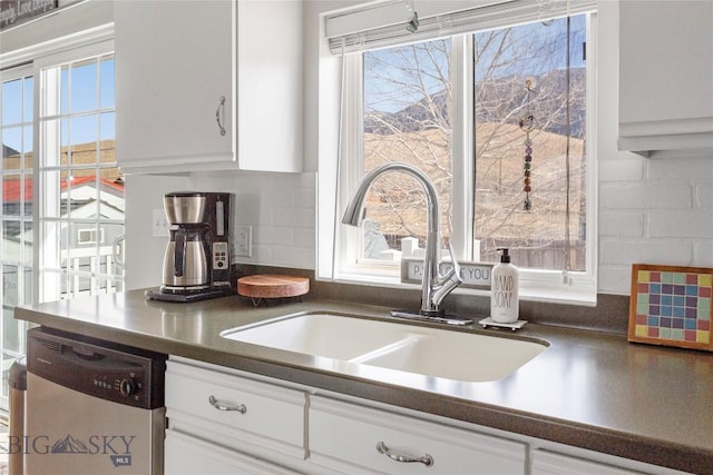 kitchen featuring decorative backsplash, dark countertops, stainless steel dishwasher, a sink, and white cabinetry
