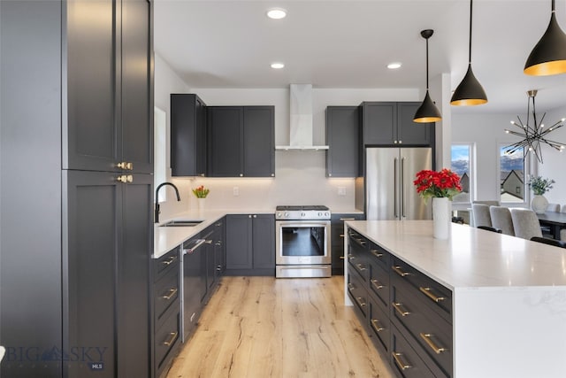 kitchen featuring appliances with stainless steel finishes, an inviting chandelier, a sink, wall chimney range hood, and light wood-type flooring