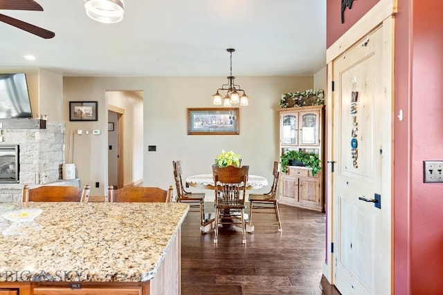 kitchen with light stone countertops, hanging light fixtures, a stone fireplace, ceiling fan with notable chandelier, and dark wood-type flooring