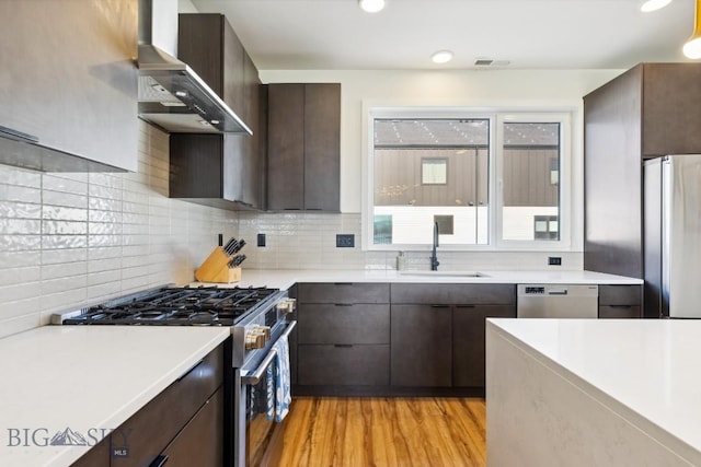 kitchen with light wood finished floors, light countertops, a sink, stainless steel appliances, and wall chimney range hood