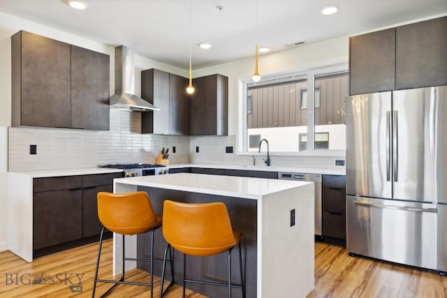 kitchen with a breakfast bar area, wall chimney exhaust hood, dark brown cabinetry, appliances with stainless steel finishes, and a kitchen island