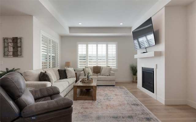 living area with recessed lighting, light wood-style flooring, a tray ceiling, baseboards, and a glass covered fireplace