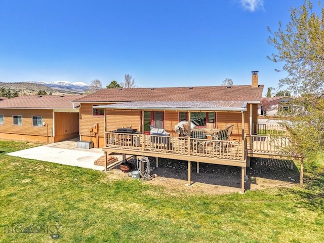 rear view of property with a yard, a chimney, a deck with mountain view, a patio, and roof with shingles