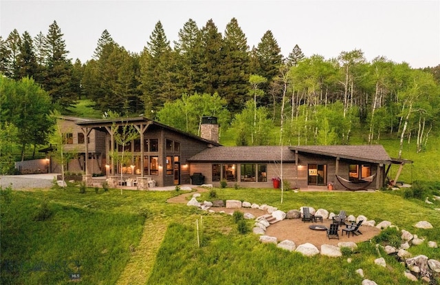 back of house featuring a fire pit, a chimney, a yard, a patio, and a wooded view