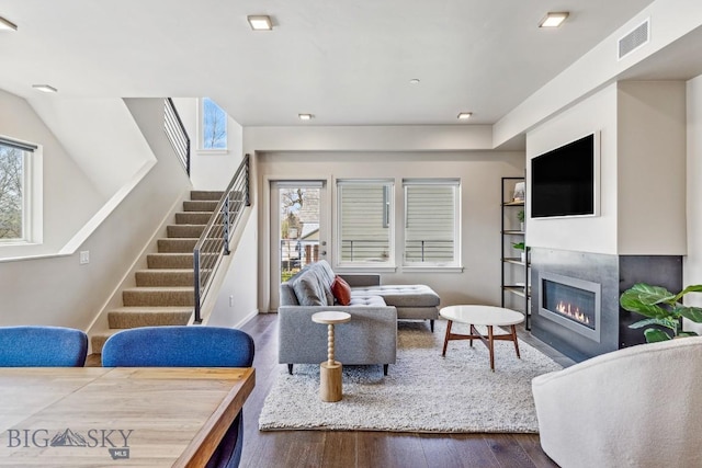 living room featuring healthy amount of natural light, wood finished floors, stairs, a glass covered fireplace, and recessed lighting