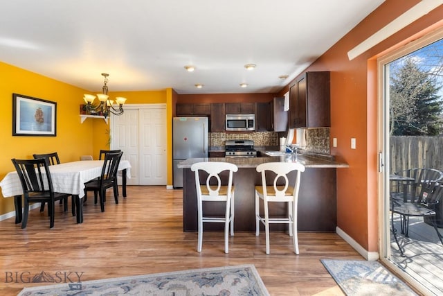 kitchen with appliances with stainless steel finishes, a chandelier, light wood-style flooring, a peninsula, and tasteful backsplash