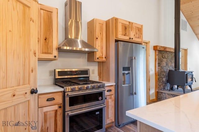 kitchen with stainless steel appliances, wall chimney range hood, a wood stove, and light brown cabinets