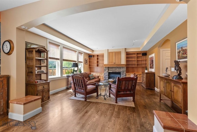 living area featuring arched walkways, baseboards, dark wood-style floors, a fireplace, and built in shelves