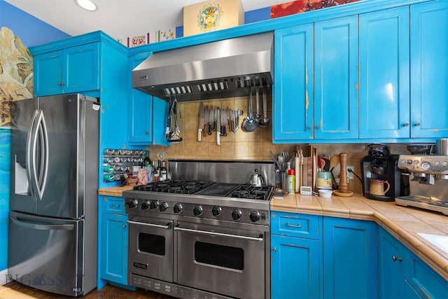 kitchen featuring appliances with stainless steel finishes, wall chimney range hood, blue cabinetry, tile countertops, and tasteful backsplash